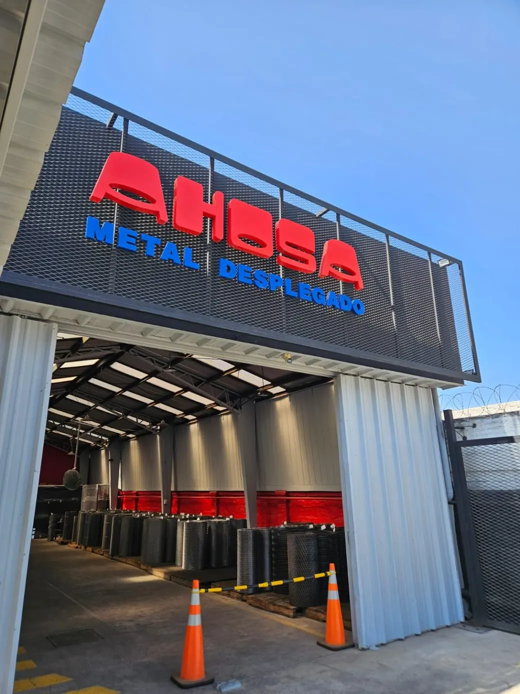 Acceso a la planta de AHOSA en Macul: el letrero AHOSA Metal Desplegado montado sobre un panel de metal desplegado y, al fondo, rollos de malla apilados en la bodega.
