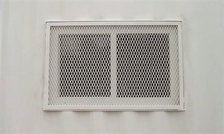 Rejilla de ventilación de metal desplegado enmarcada e instalada en el muro de un edificio, pintada de blanco.